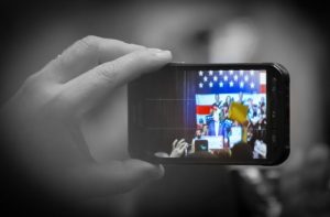 A black and white photo of hand holding up a cell phone camera to photograph President Trump