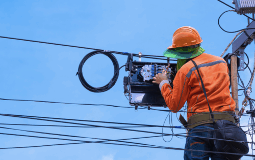 A lineman is suspended in air working inside a fiber splice case amid utility lines