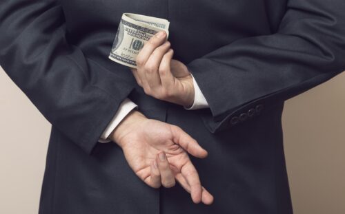 Businessman in a suit holding his hands behind his back with stack of dollar banknotes in one and fingers crossed on another one