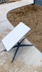 A Starlink dish sits on a concrete walkway with a wire connecting to home in background