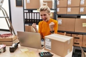 A business owner pumps her fist while looking at her laptop inside a warehouse with dozens of cardboard boxes
