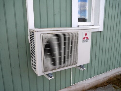 White heat pump condenser unit mounted on wall brackets against green siding, below a window.