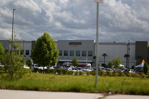 An Amazon warehouse near a busy parking lot under a cloudy sky. Sellers say Amazon's monopoly is getting worse.