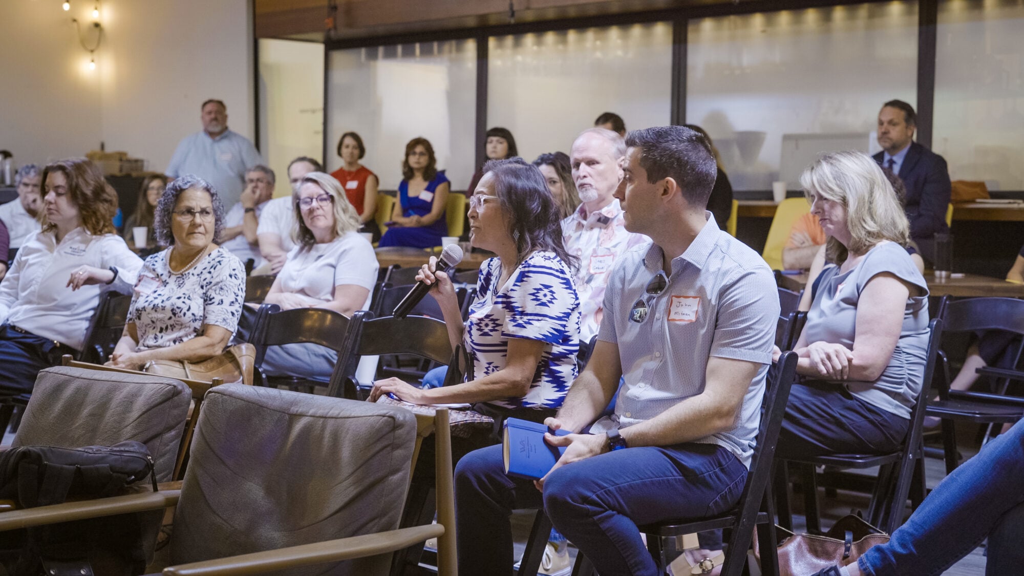 Several people sit in a crowd, one of whom is asking a question into a microphone