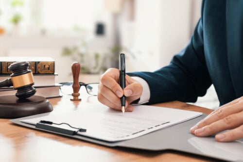 Someone in a suit signing a piece of paper on a desk with a gavel and stamp on it